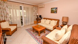 A living room with a sofa, chairs, a coffee table, and a TV. There is a window with curtains, an air conditioning unit, and a painting on the wall. - Charming Dominicus Bayahibe Apartment 2 bedrooms (Dominicus, Bayahibe)