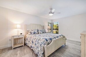 Une chambre à coucher comprenant un lit, une table de chevet, une lampe, un ventilateur de plafond et une fenêtre avec des stores.