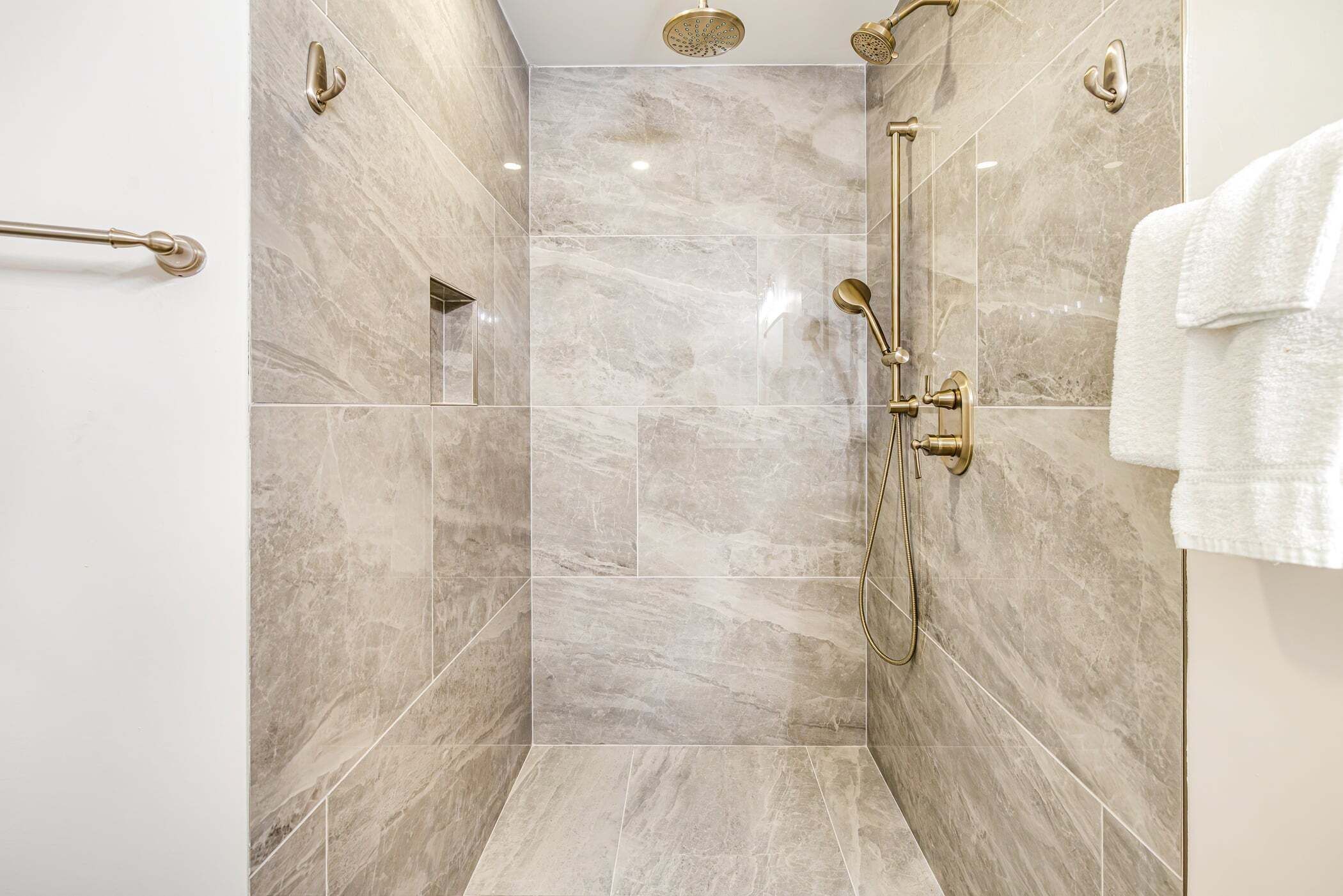 A modern shower with a handheld showerhead and a wall-mounted rain showerhead, marble tiles, and a towel rack.