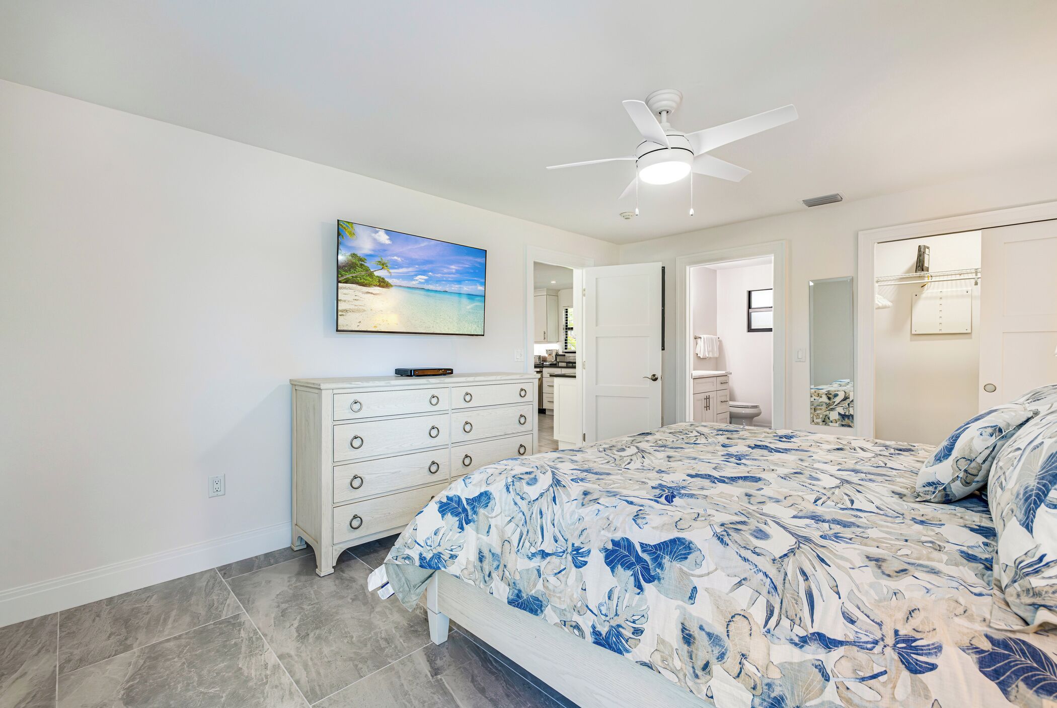 Une chambre à coucher avec un lit, une commode, un ventilateur de plafond, un tableau et une salle de bain visible à travers une porte ouverte.