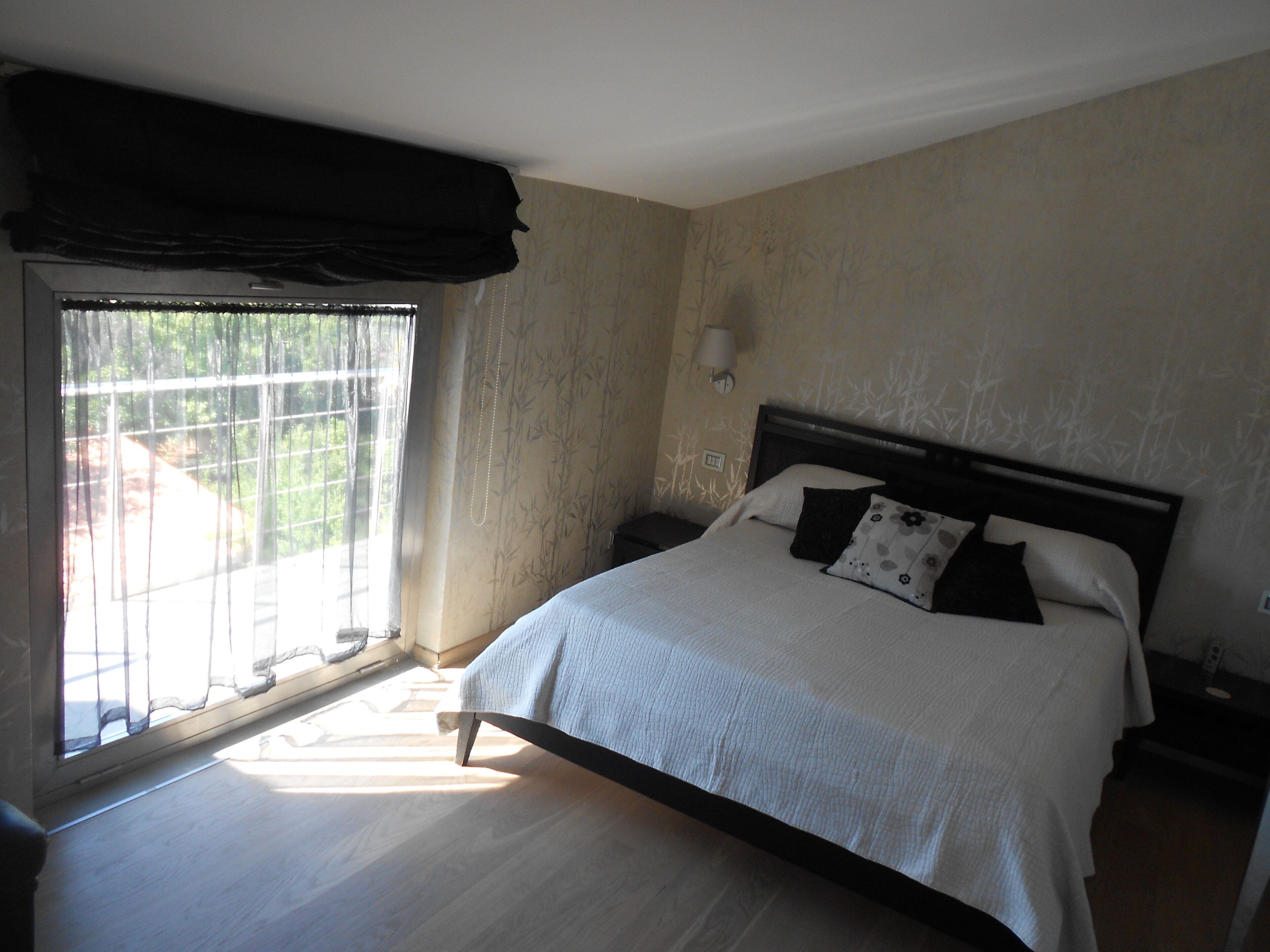 Un dormitorio con cama, mesita de noche, lámpara de pared y ventana con cortinas.