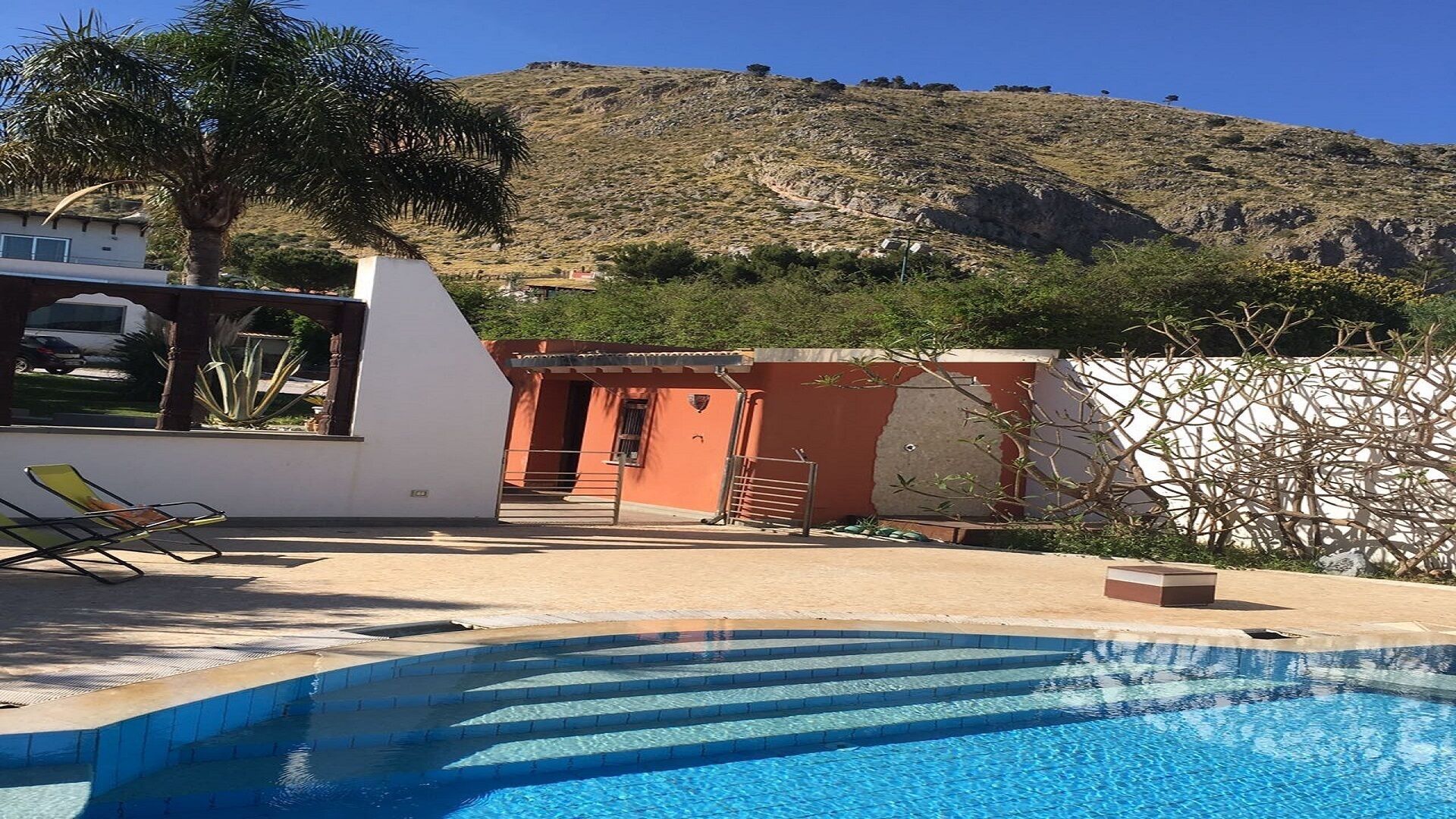 Une piscine avec des marches menant à l’intérieur, un bâtiment blanc avec une porte rouge et une colline en arrière-plan.