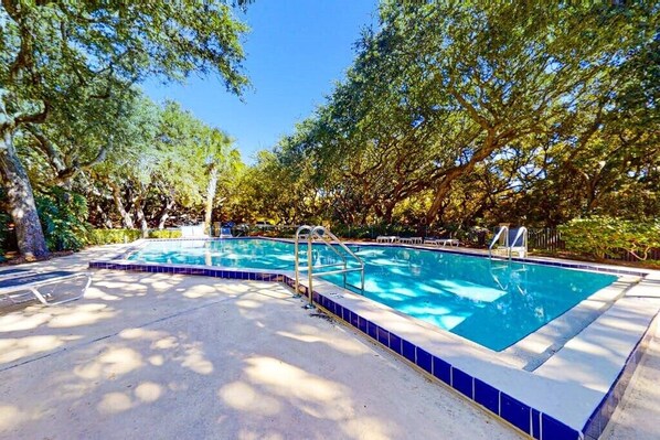 Piscina rectangular con azulejos azules, rodeada de árboles y un solárium de hormigón.