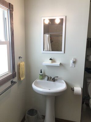 Une salle de bain avec un lavabo blanc, un miroir avec éclairage, un porte-serviettes, une petite étagère et une poubelle.