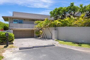 A two-story residential building with a garage, a driveway, and a small garden area with palm trees.