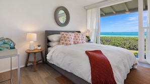 A bedroom with a bed, a nightstand, a lamp, a mirror, and a view of the ocean. - Pueohala: Spectacular Kailua Beachfront - updated Beach House (Kailua)