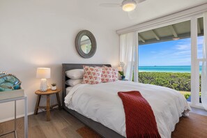 A bedroom with a bed, a nightstand, a lamp, a mirror, and a view of the ocean.