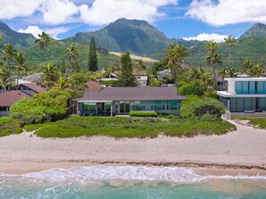 A beachfront property with a modern house, lush greenery, and a mountain backdrop.
