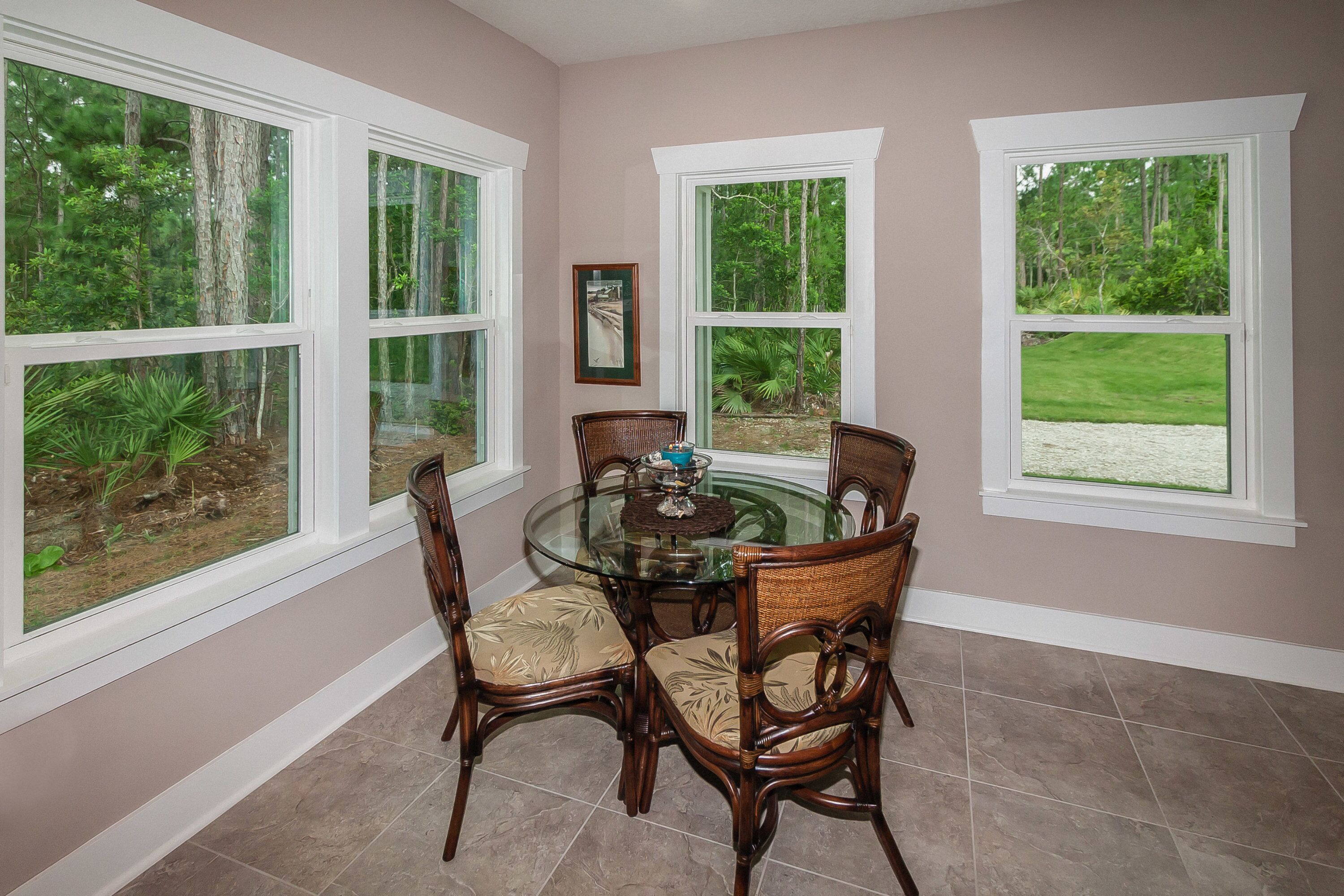 A dining area with a glass table surrounded by wicker chairs, large windows with a view of greenery, and a framed picture on the wall.