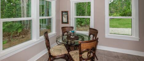 A dining area with a glass table surrounded by wicker chairs, large windows with a view of greenery, and a framed picture on the wall.