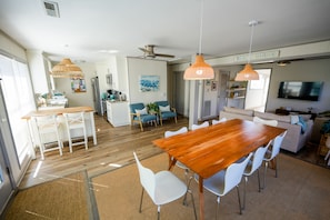 Uma sala de estar ampla com uma mesa de jantar de madeira, cadeiras brancas, um sofá e uma televisão.