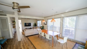 A spacious living area with a wooden dining table, white chairs, a ceiling fan, and large windows with blinds.
