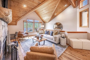 A cozy loft-style living room with a high ceiling, wooden beams, a fireplace, and a large window offering a view of the outdoors.