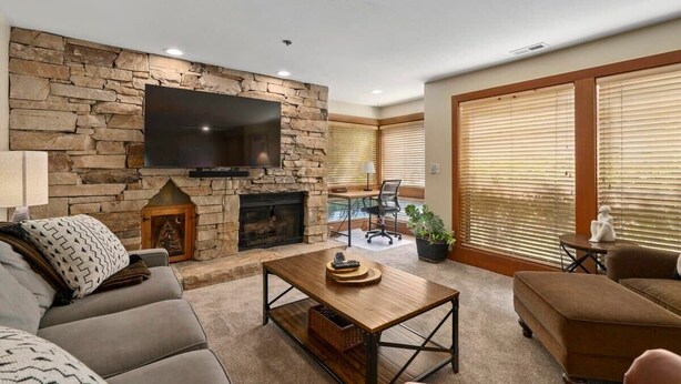 A living room with a stone wall, a flat-screen TV, a fireplace, and a large window with blinds. - The Silver Cliff, 2BR Walk to Lifts and Main St! (Park City)