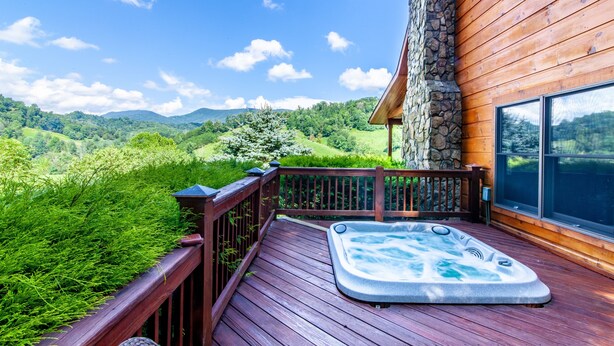 Una terraza de madera con un jacuzzi y un muro de piedra, con vistas a un paisaje verde y exuberante.