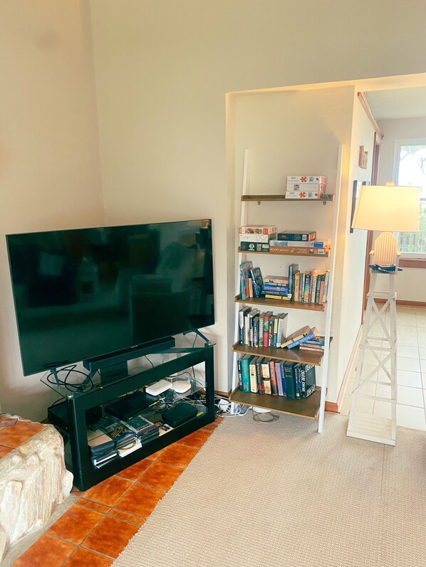 A living room with a flat-screen TV on a stand, a bookshelf filled with books and boxes, a lamp, and a small table.