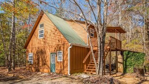 A wooden cabin with a green roof, a porch, and a staircase leading to a deck.