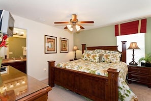 A bedroom with a wooden bed, a ceiling fan, two framed pictures, a bedside table with a lamp, and a window with red curtains. - Ly230329 - Arcadia Estates - 7 Bed 6.5 Baths Villa (Kissimmee)