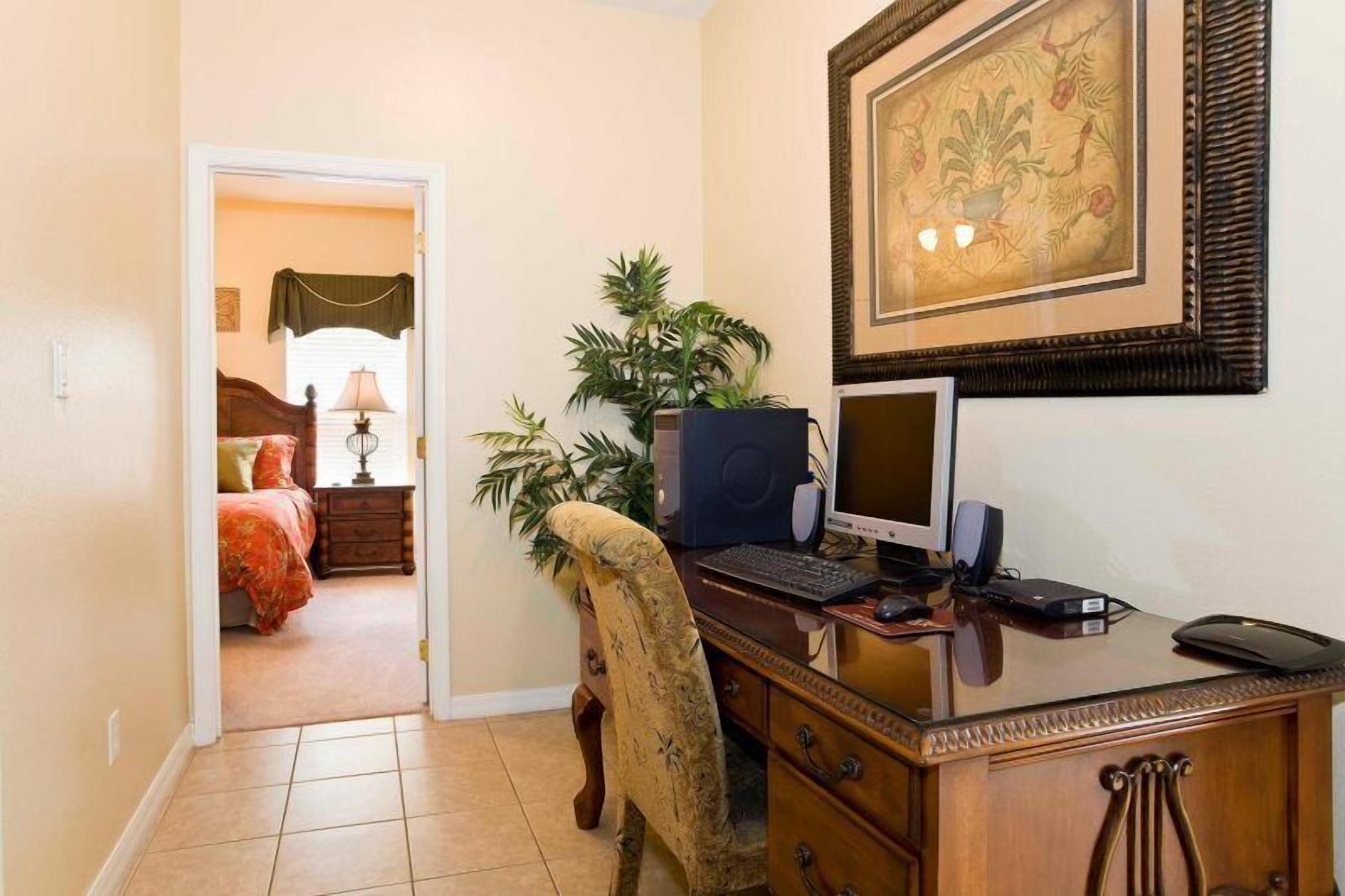 A bedroom with a bed, a nightstand, and a lamp. There is a desk with a computer, a chair, and a plant.