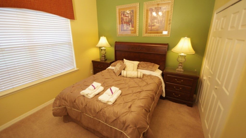 A bedroom with a bed, two bedside tables with lamps, a window with blinds, and two framed pictures on the wall.