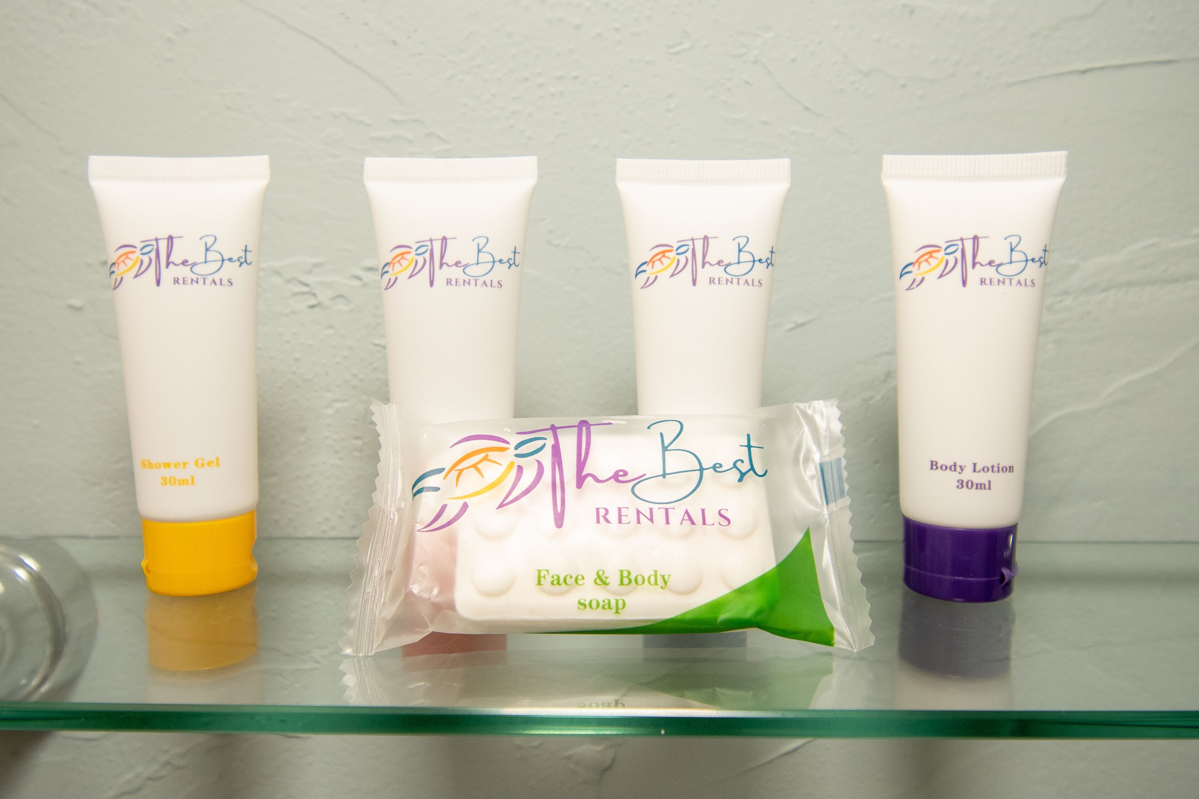 Un estante de baño con cuatro artículos de aseo: un tubo amarillo de gel de ducha, un tubo blanco de loción corporal, un tubo blanco de jabón facial y corporal, y un tubo morado de loción corporal.