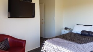 A hotel room with a bed, a red chair, a nightstand, a TV mounted on the wall, and a door leading to another room. - Annandale Accommodation (Taupo)