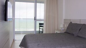 Une chambre d’hôtel avec un lit, une télévision, une chaise, une table de chevet et une vue sur la mer, visible à travers une grande fenêtre.