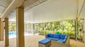 A covered outdoor area with a wicker sofa and ottoman, large windows, and a pool in the background.