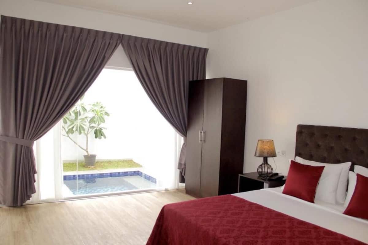 Ein Hotelzimmer mit Bett, Nachttisch, Lampe, Kleiderschrank und Blick auf Pool und Palmen.