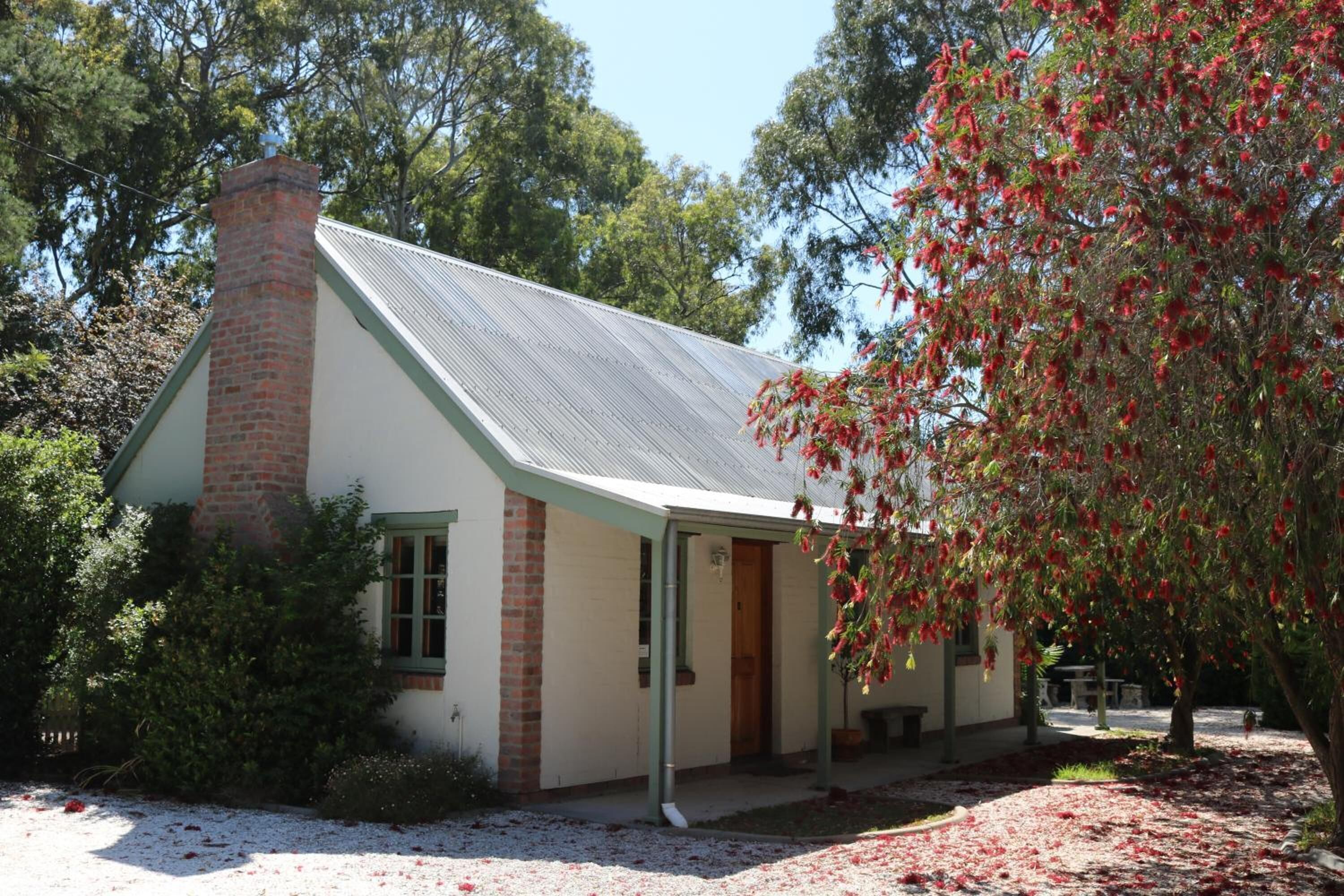 A small, single-story building with a brick chimney, a covered porch, and a garden area with red flowering plants.