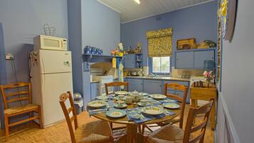 A dining area with a round table set for four, surrounded by wooden chairs. A refrigerator and microwave are visible in the background.