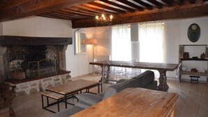 A rustic interior with exposed wooden beams, a stone fireplace, a wooden dining table, and clear plastic chairs.