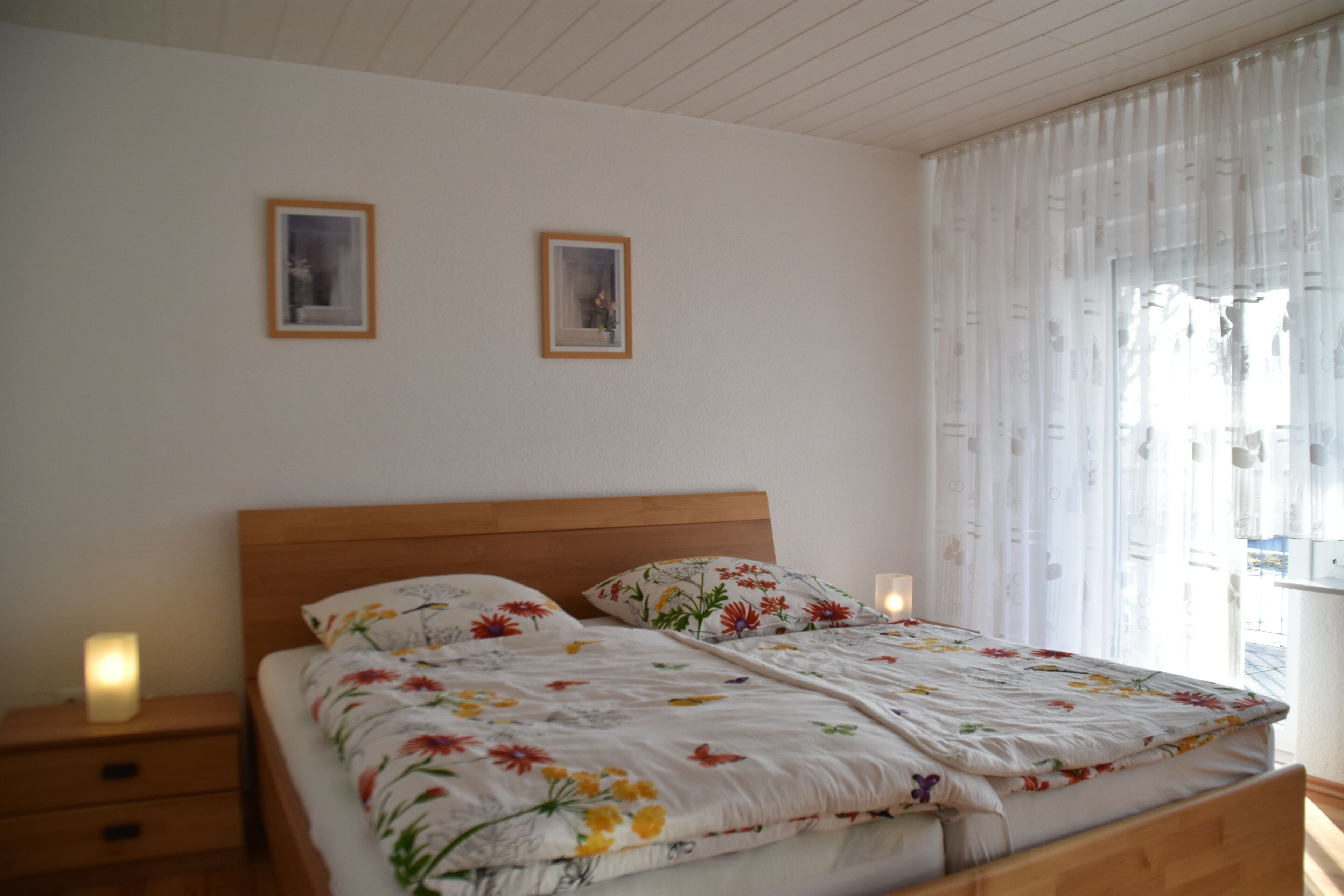 A bedroom with a wooden bed, floral bedding, two framed pictures on the wall, and a window with sheer curtains.