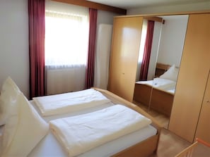 A hotel room with two beds, a wardrobe, a mirror, and a window with curtains. - Vacation apartment B, 4 persons with balcony or terrace, bedroom, 50 to 55 sqm (Inzell)