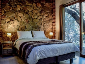 A bedroom with a stone wall, a bed with white linens, two bedside lamps, and a large window with a view of trees. - Cabin between Forest and River (Lo Barnechea)