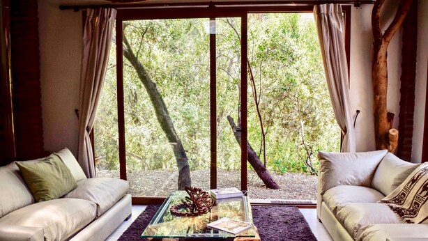 A room with two sofas facing a large window with a view of trees. A glass coffee table with decorative items is in the center. - Cabin between Forest and River  (Lo Barnechea)