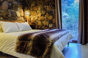 Un dormitorio con pared de piedra, cama con ropa de cama blanca y vista a zonas verdes.