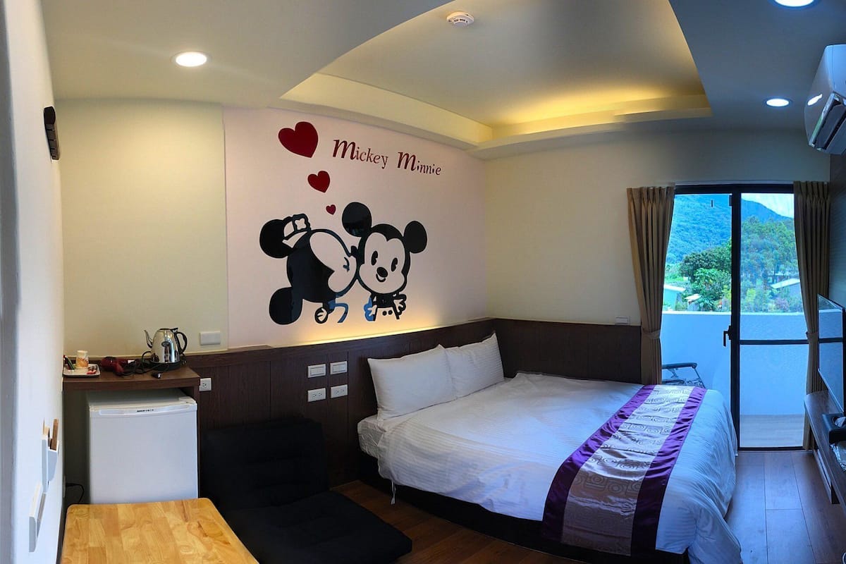 Quarto de hotel com cama, pequena escrivaninha, chaleira e janela com vista para o verde.