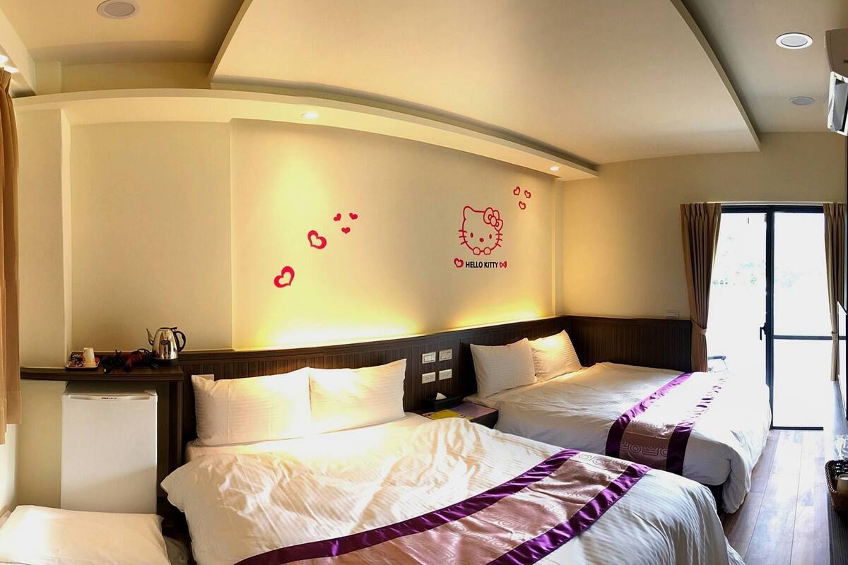 Quarto de hotel com duas camas, uma pequena escrivaninha com chaleira, parede decorada com tema Hello Kitty e uma janela com cortinas.