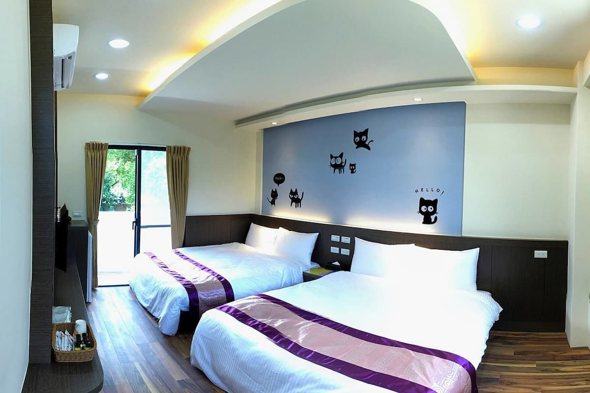 Quarto de hotel com duas camas, uma janela grande com cortinas, parede com adesivos de gato e uma lâmpada no teto.