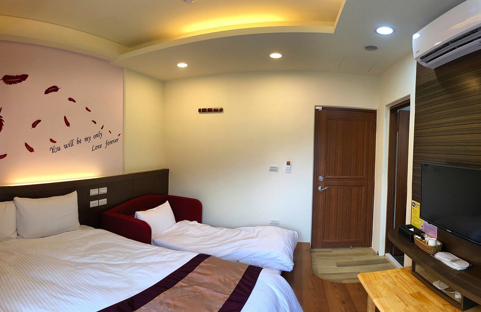 Habitación de hotel con una cama grande, una cama más pequeña, un televisor, una puerta de madera y una pared con una cita romántica.
