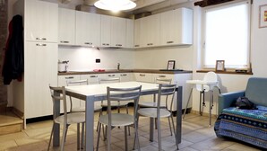Una cocina con armarios blancos, una mesa de comedor con cuatro sillas, un ventanal con vistas y un sofá azul con una manta estampada.