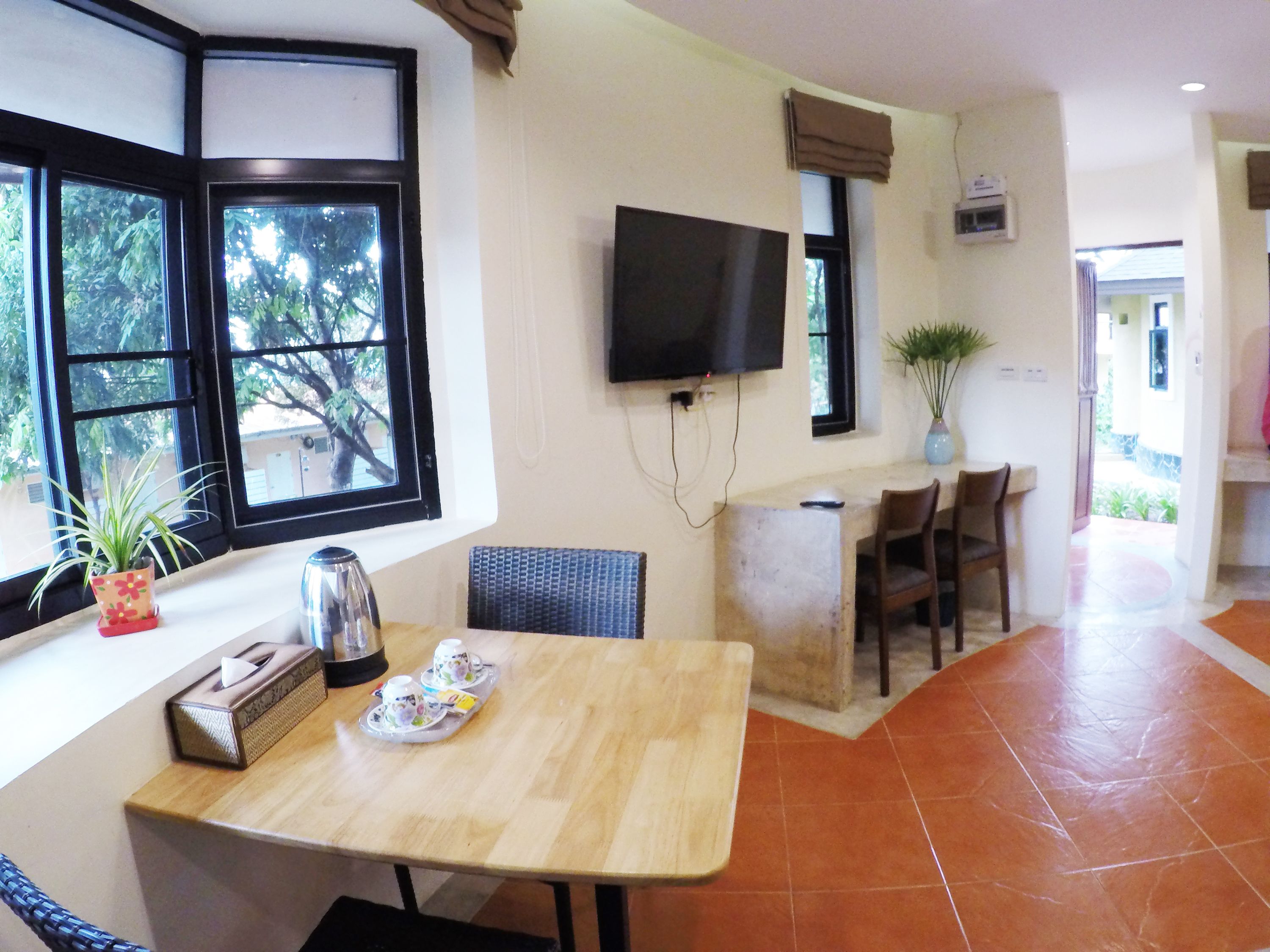 A room with a wooden table, chairs, a TV mounted on the wall, a window with a view of trees, and a potted plant.