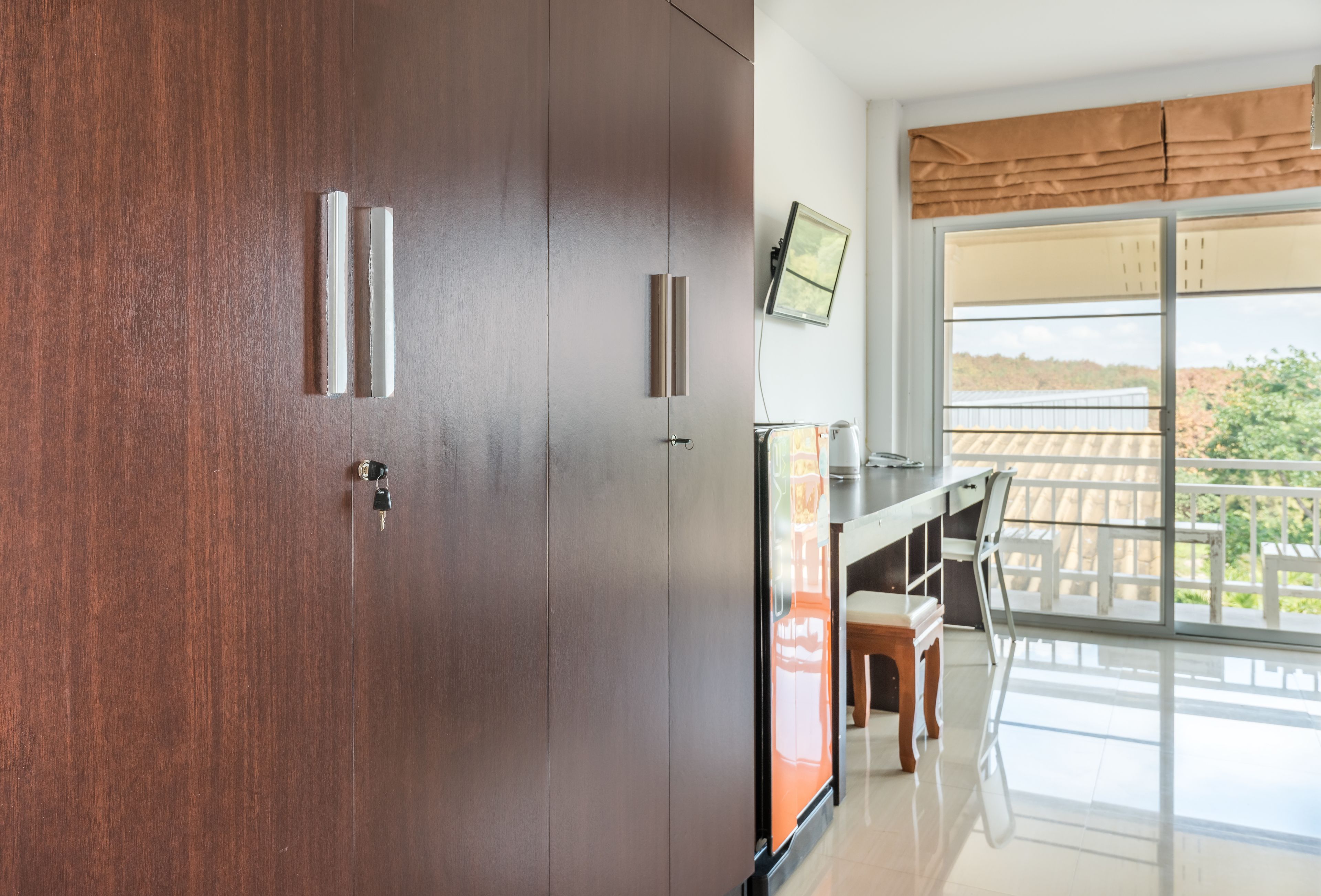 A modern room with a wooden door, a flat-screen TV mounted on the wall, a dining area with a table and chairs, and a balcony with a view of greenery.