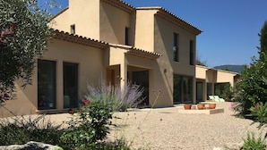 Ein modernes, zweistöckiges Haus mit großen Fenstern, einer Terrasse mit Gartenmöbeln und einem angelegten Garten mit verschiedenen Pflanzen und einem Felsenelement.