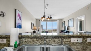 A kitchen with a granite countertop, stainless steel sink, and a view of the ocean through large windows.