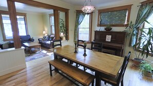 Un coin repas avec une table en bois, des bancs et des chaises. Un lustre est suspendu au-dessus de la table. La pièce est dotée de grandes fenêtres avec des rideaux, et on peut apercevoir un piano au fond.