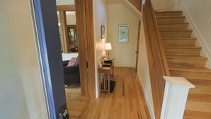 Un escalier en bois menant à un deuxième étage, un couloir avec une lampe et une petite table, et une porte donnant sur un espace de vie.