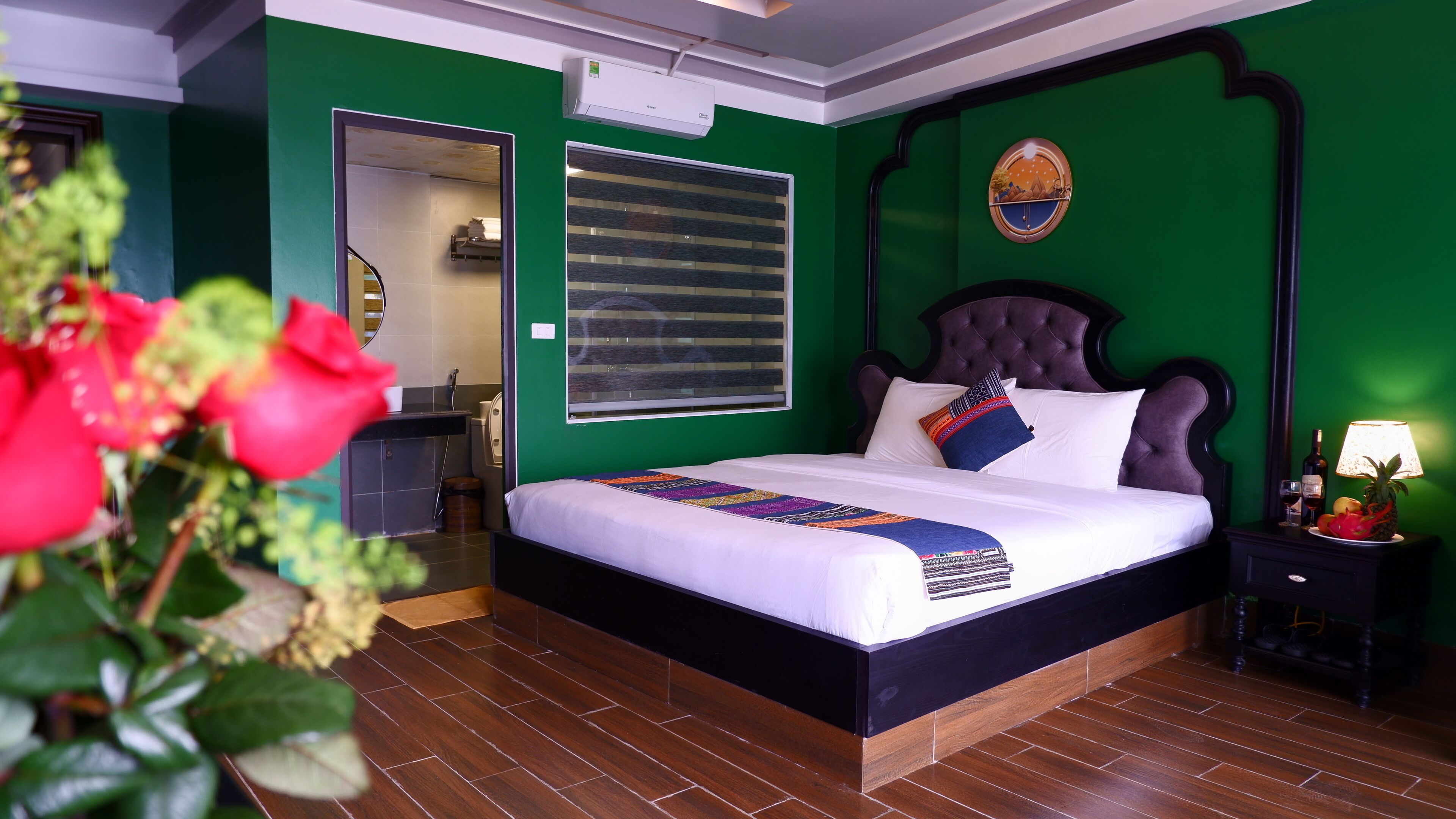 A hotel room with a large bed, a nightstand with a lamp, a window with blinds, and a green wall.