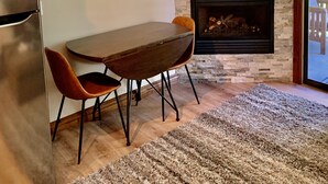 Un área de comedor con una mesa y sillas, una chimenea, un televisor montado encima y una zona de cocina con un refrigerador.